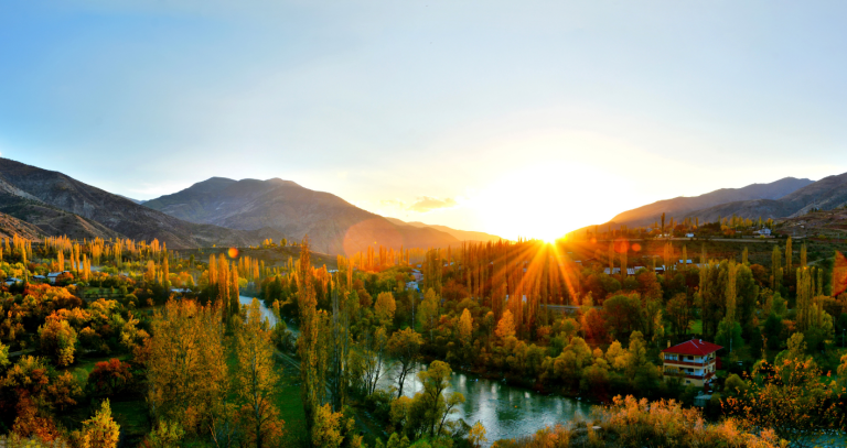The sun peaks out with golden rays over a mountain and river through the trees.